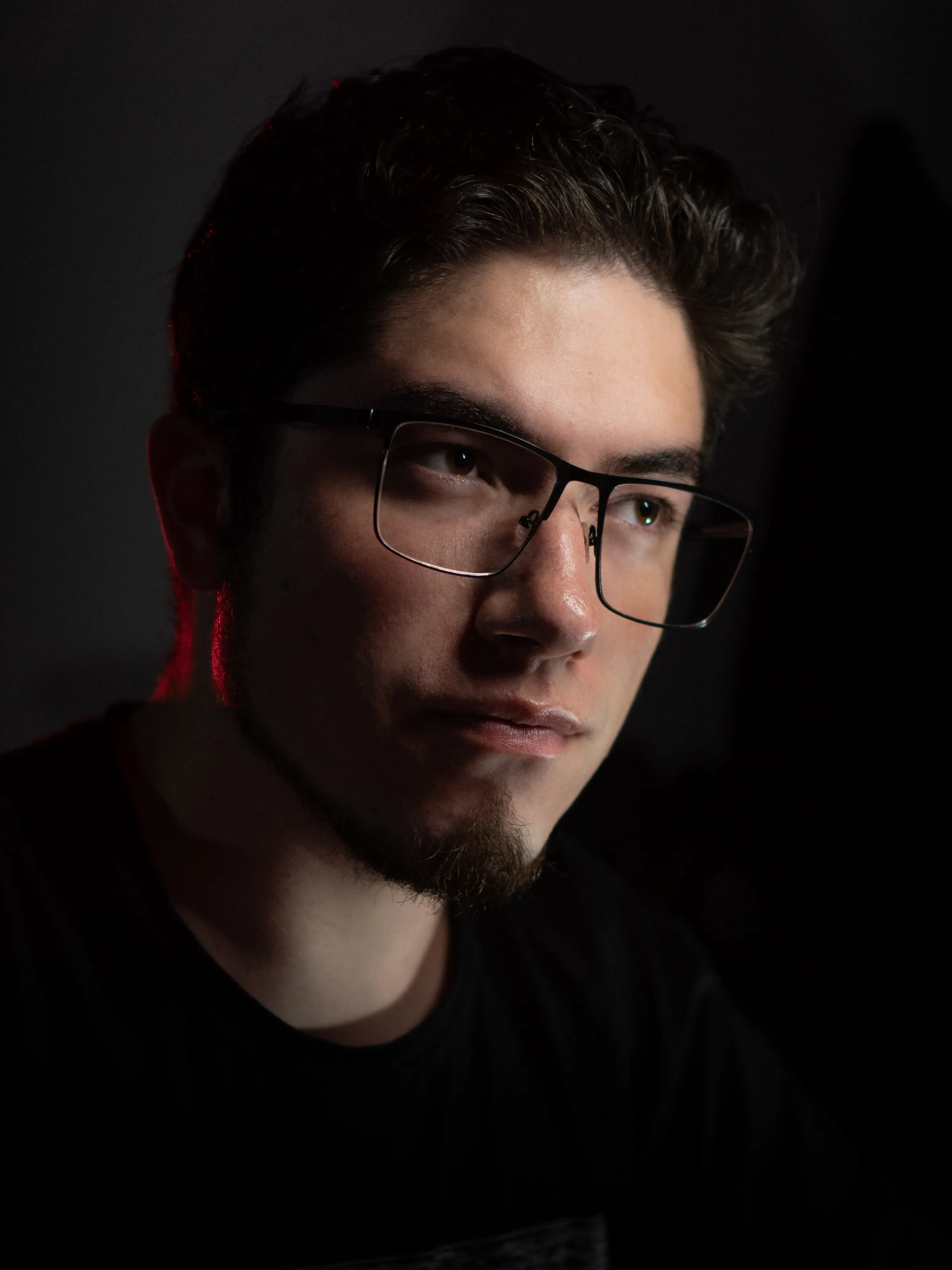 A man with glasses emerges from a dark background in side light.