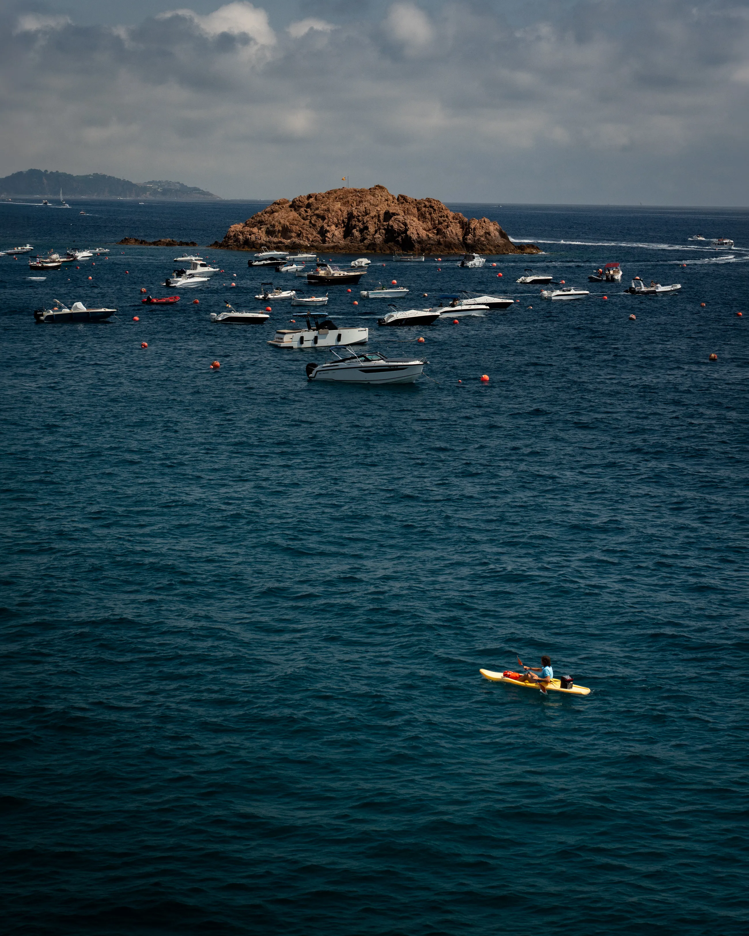 A person in a yellow kayak paddles past anchored boats and a rocky islet off Lloret de Mar.
