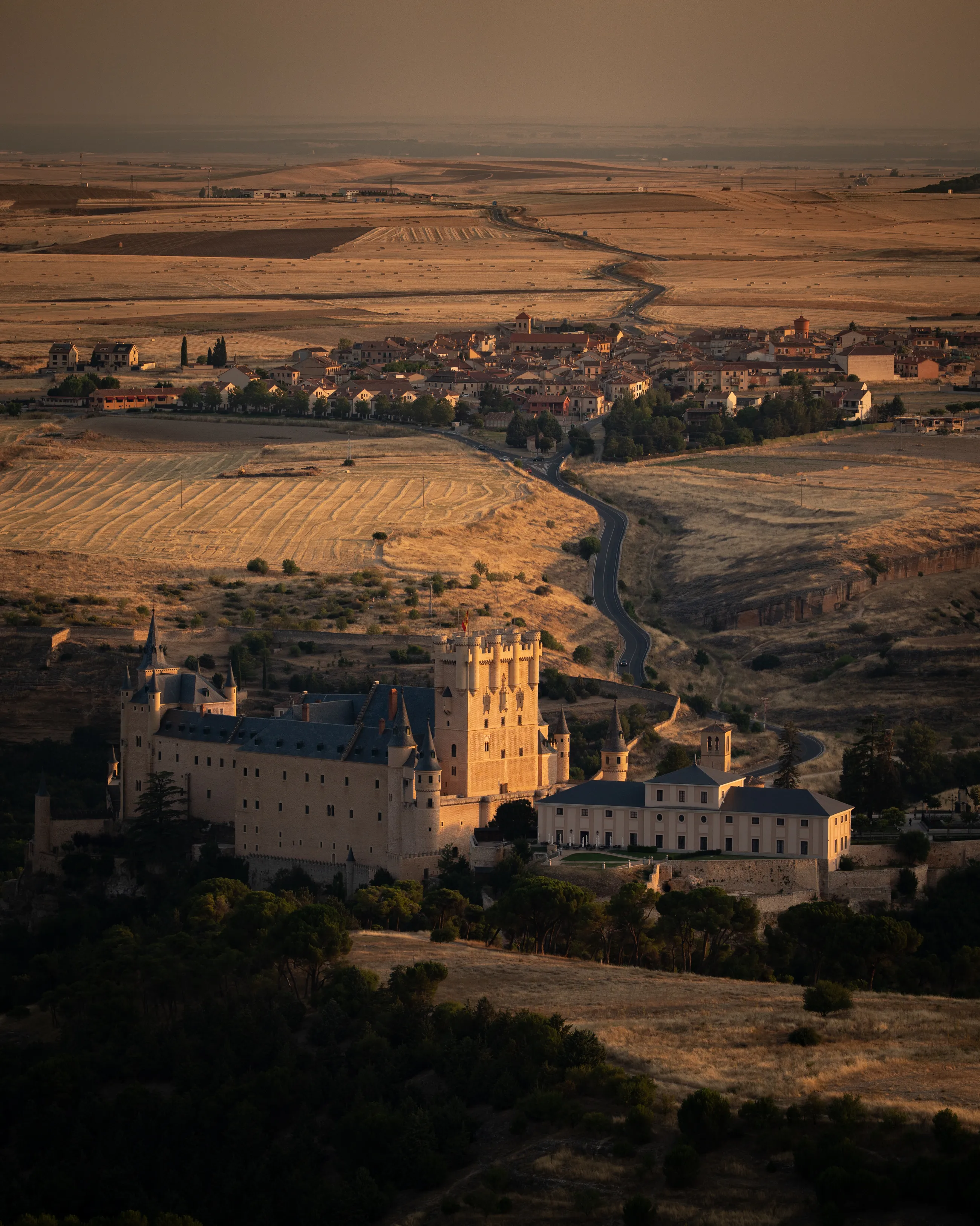 The Alcázar of Segovia catches warm evening light beneath a village and golden fields.