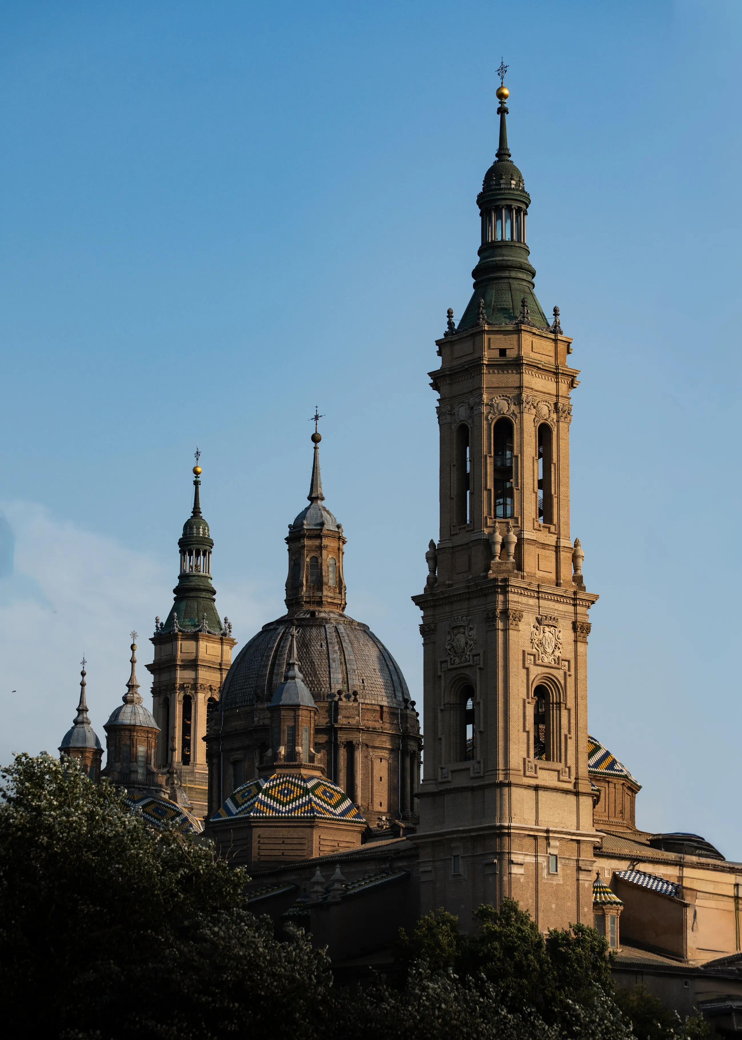 Sunlit towers and patterned tiled domes rise above trees against a blue sky in Zaragoza.
